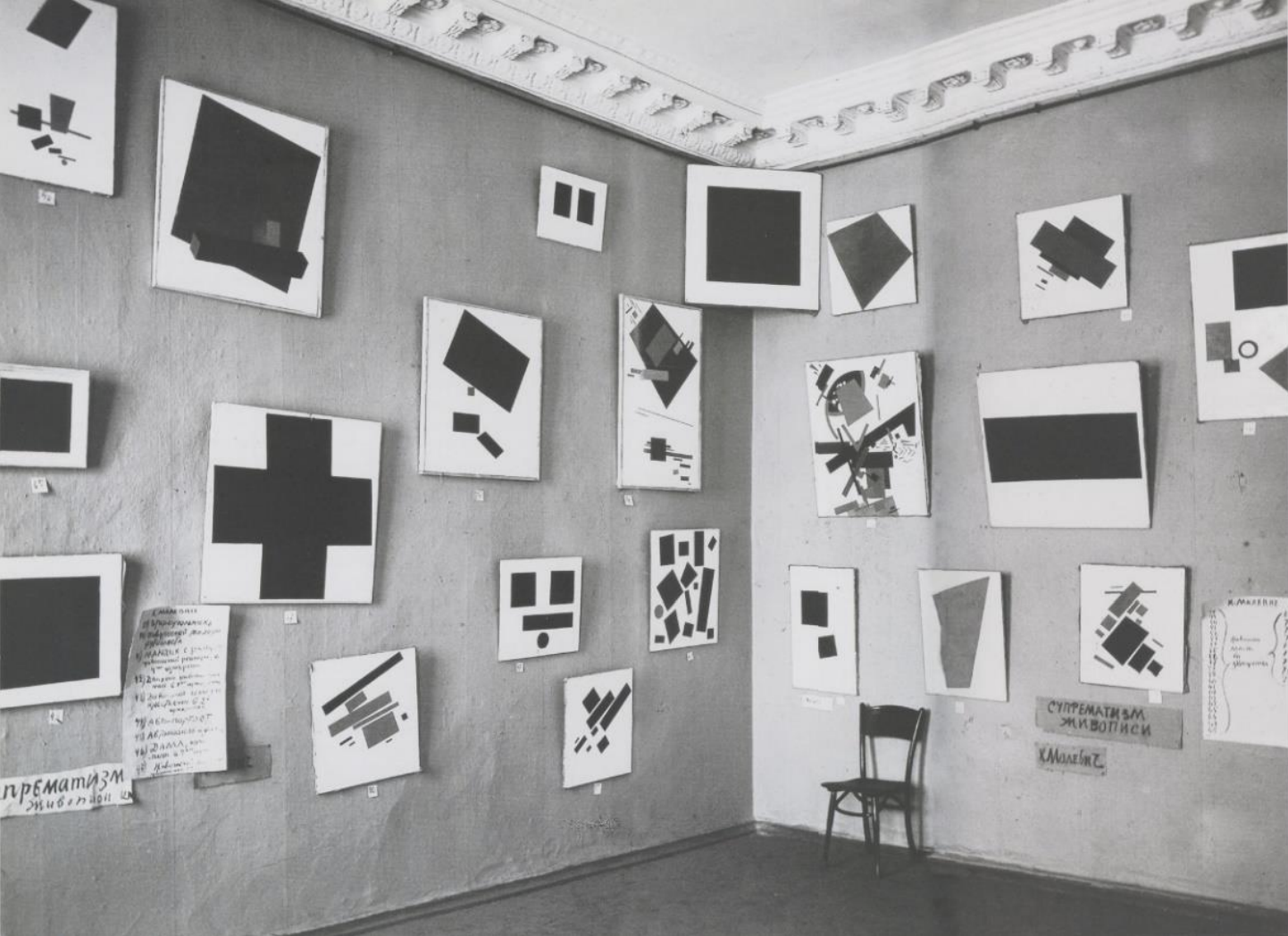 Schwarz-weiße Ausstellung mit abstrakten geometrischen Gemälden an einer Wand und einem Stuhl.